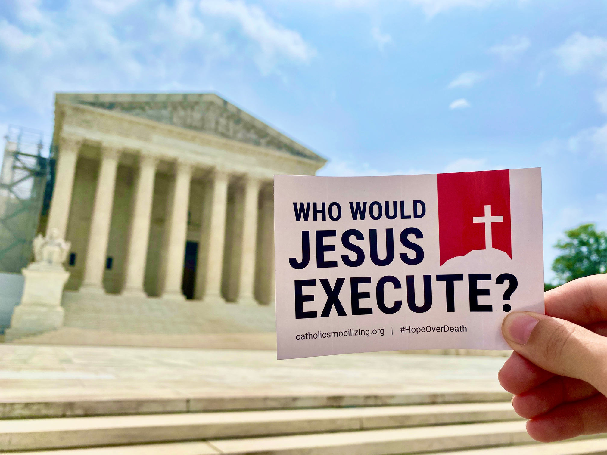 Hand holds sticker that says "Who would jesus execute?" in front of the U.S. Supreme Court